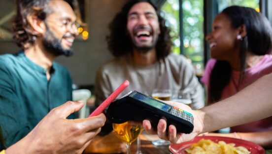 Stock image of happy friends using nfc technology on smartphone to pay for their meal at a restaurant, enjoying drinks and food.