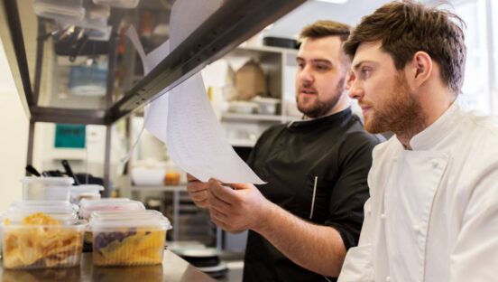 Chef and cook with lists or bills at kitchen.