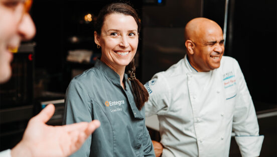 Image of Chef Lynn Hay from Entegra's Culinary Consulting, in a grey chef coat, speaking with 2 associates.